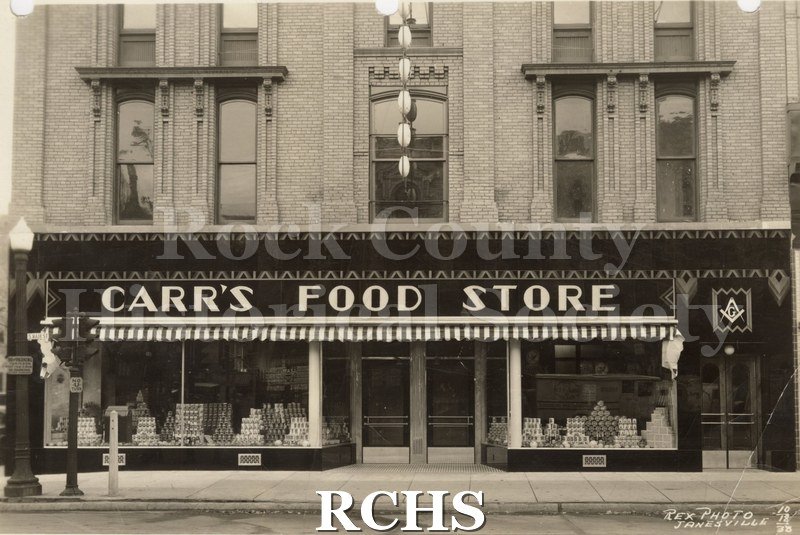 Black and white photograph of exterior of Carr's Food Store. Shows displays of foods in the window. Photographer: Rex Photo Janesville 10-18-33.
