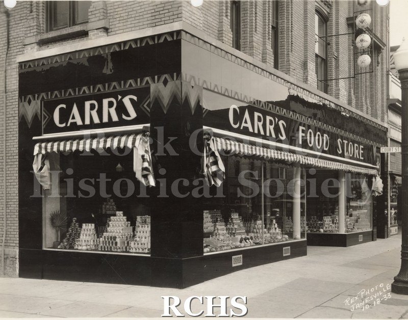 Black and white photograph of the exterior of Carr's Food Store. Shows displays of foods in the window. Photographer: Rex Photo Janesville 10-18-33.