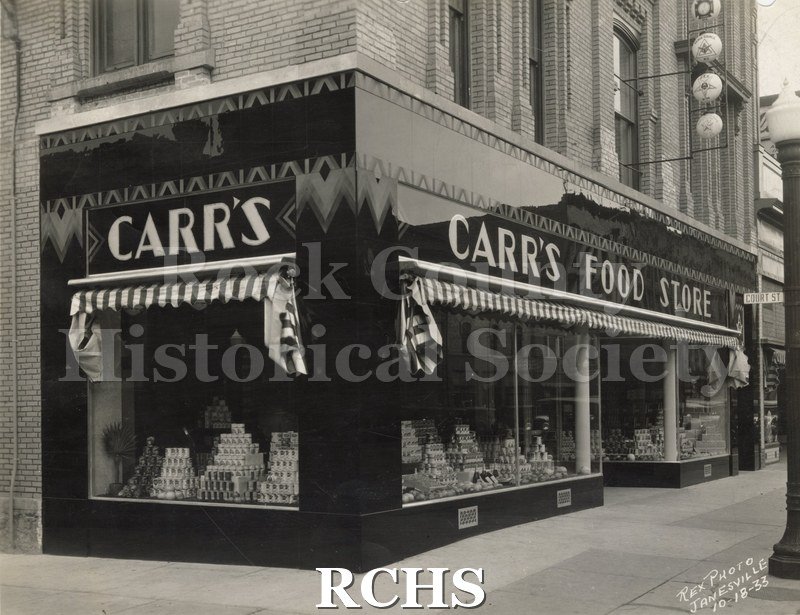 Black and white photograph of the exterior of Carr's Food Store. Shows displays of foods in the window. Photographer: Rex Photo Janesville 10-18-33.