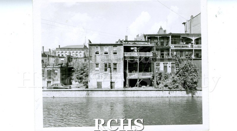 Black and white photograph of 13 N. Main St. Shows the back side of stores on Main Street. The river is in the foreground.