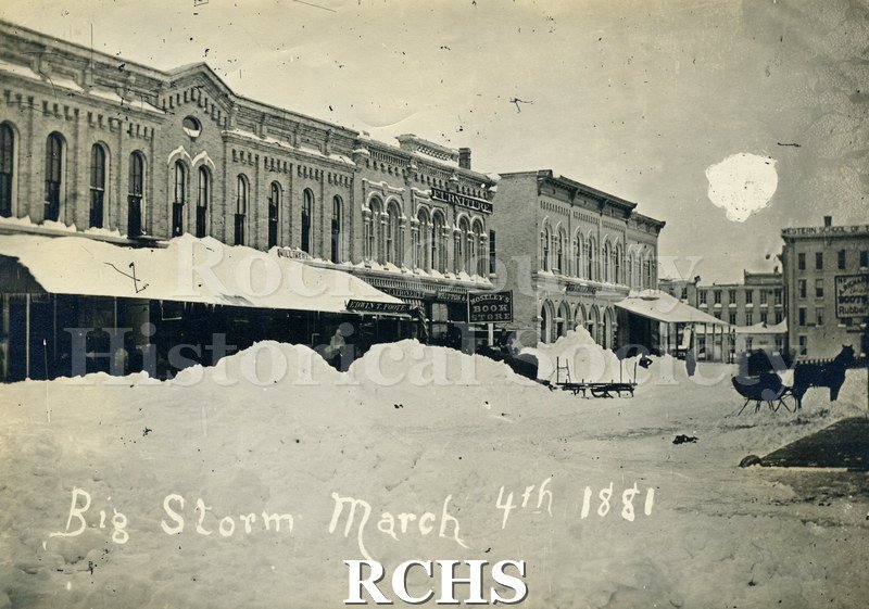 Black and white photograph of the Big Storm March 4th 1881. Shows buildings with large piles of snow in front of them; horse pulling sleigh is on the street.