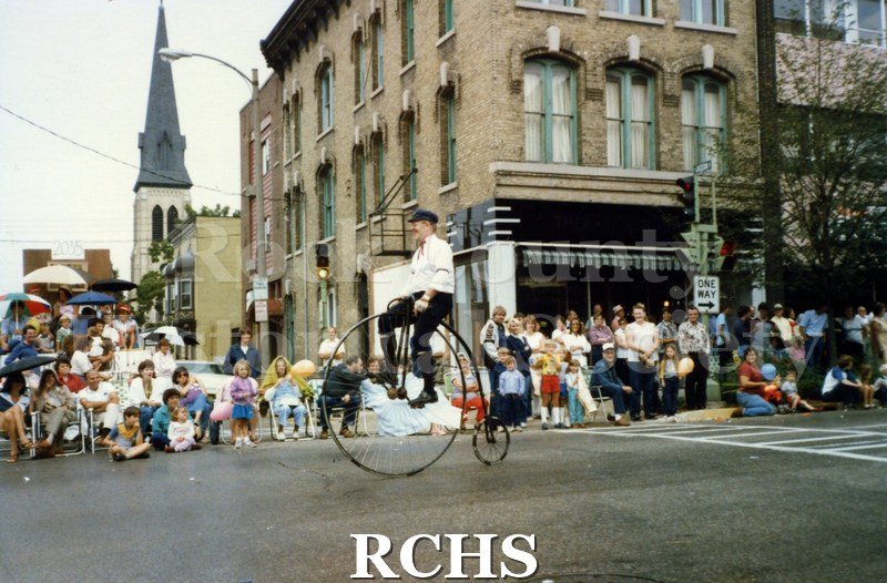 Historic image of Colored photogrpah of a man riding a penny-farthing bicycle (one very large wheel in front, one very small wheel in back, rider sits atop the.
