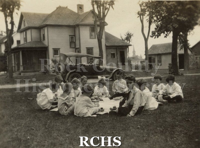 Black and white photograph of several children having a picnic. There is a two story house in the background and a car parked on the road behind the children.