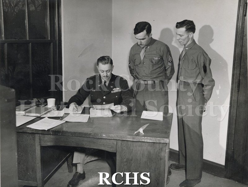 Black and white photograph of Maj. Patterson, Sgt.Bolton and John Higgins. Two servicemen are standing beside another serviceman sitting at a desk looking at papers.
