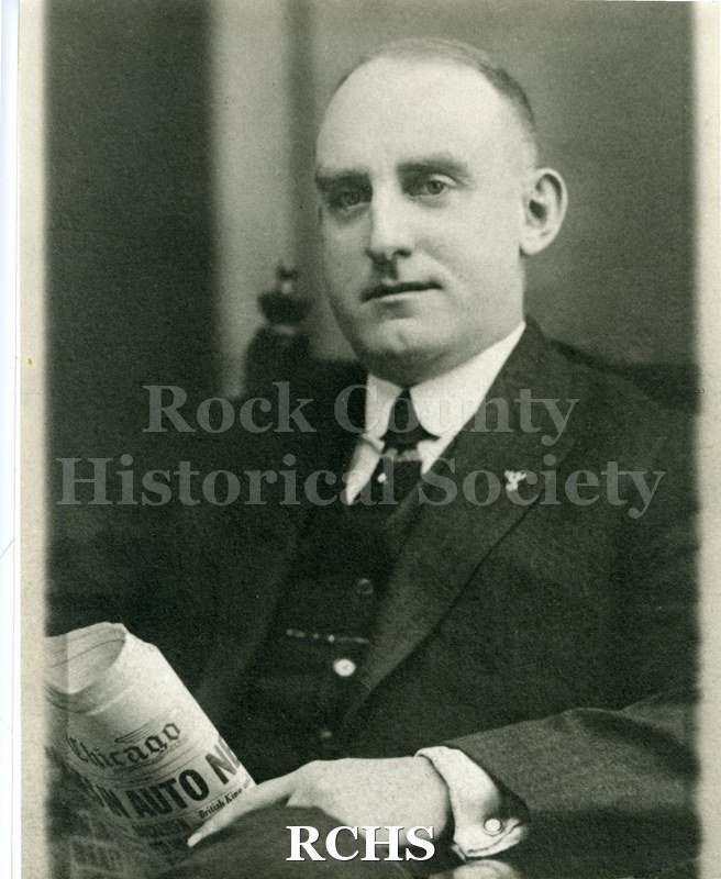 Black and white photograph of Charles Muggleton sitting in a chair. He is wearing a dark suit coat, vest, white shirt and tie and is holding a Chicago newspaper in.