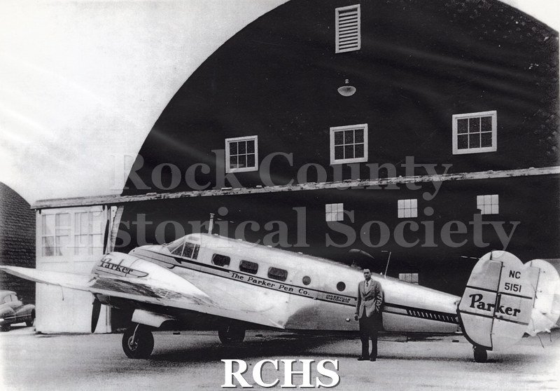 Black and white photograph of Parker Pen Co. airplane at Rock County Airport. Side view of airplane with man standing in front.