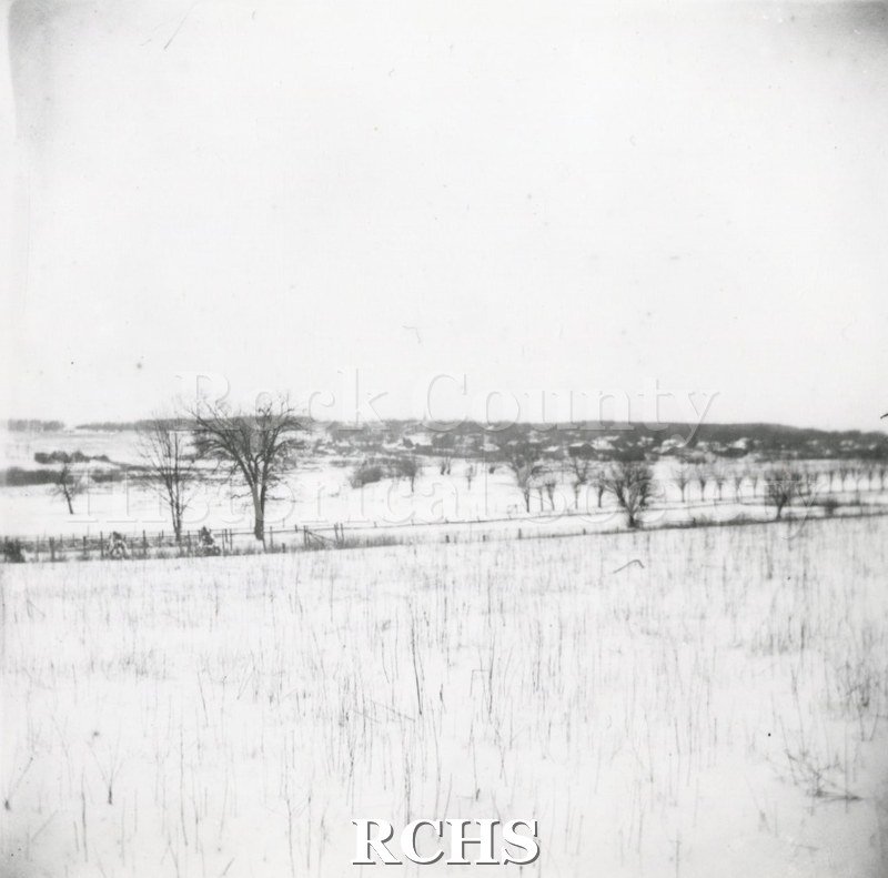 Historic image of Blackand white copy print of Shopiere farm land. Farm fields are bisected by a road with trees along it. The land is covered in snow.