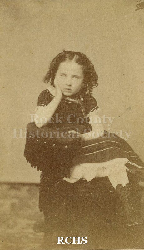 Black and white photograph of Anna Fisher. She is a younng girl sitting in a chair wearing a dark short sleeved dress.