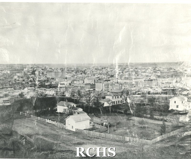 Historic image of Black and white copy print of early Janesville. Shows buildings in the background; and a farm with house and barn in the foreground.