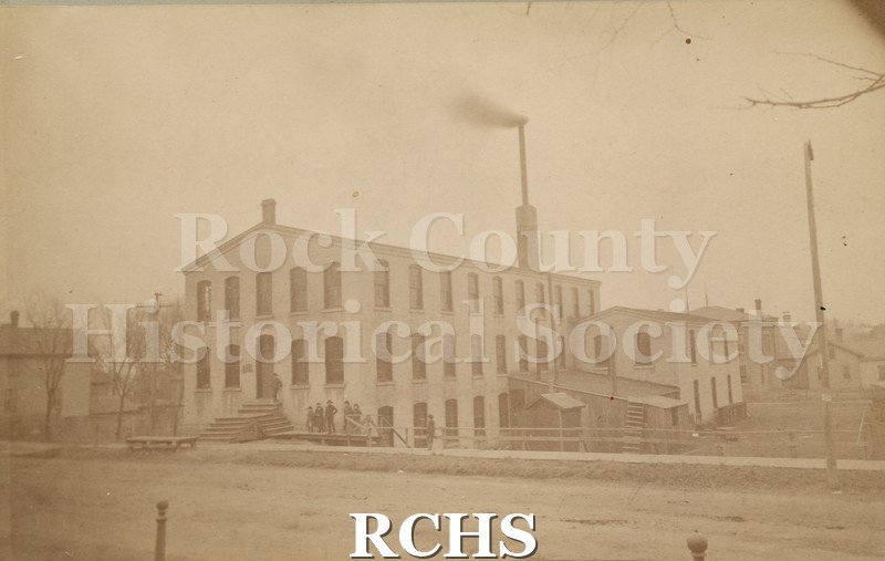 Black and white photograph of the Lewis Knitting Co. Shows a two story building with a large chimney on top.