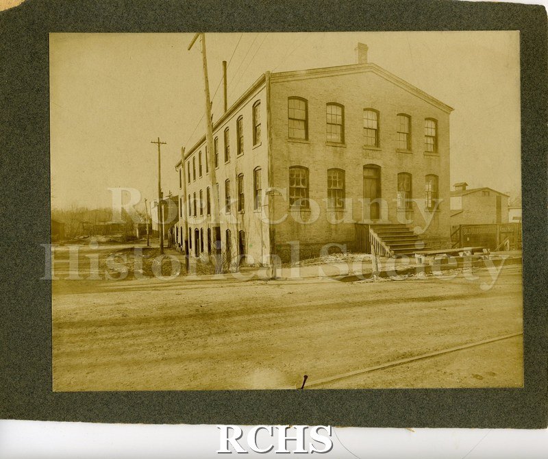 Black and white photograph of the Lewis Knitting Co. Shows a two story building with a large chimney on top. There are steps leading up to the doorway.