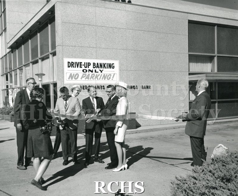 Black and white photograph of the Merchants & Savings Bank ribbon cutting ceremony. There are six men and two women standing by the ribbon.