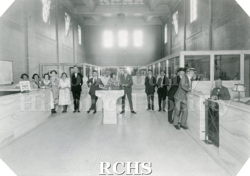 Black and white photograph of the inside of Merchants and Savings Bank showing a large number of people standing in the room.