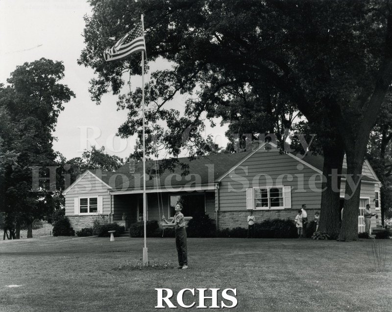 Black and white photograph of Charles Graves hoisting a flag in front of a house. There are other children and a man near the house.