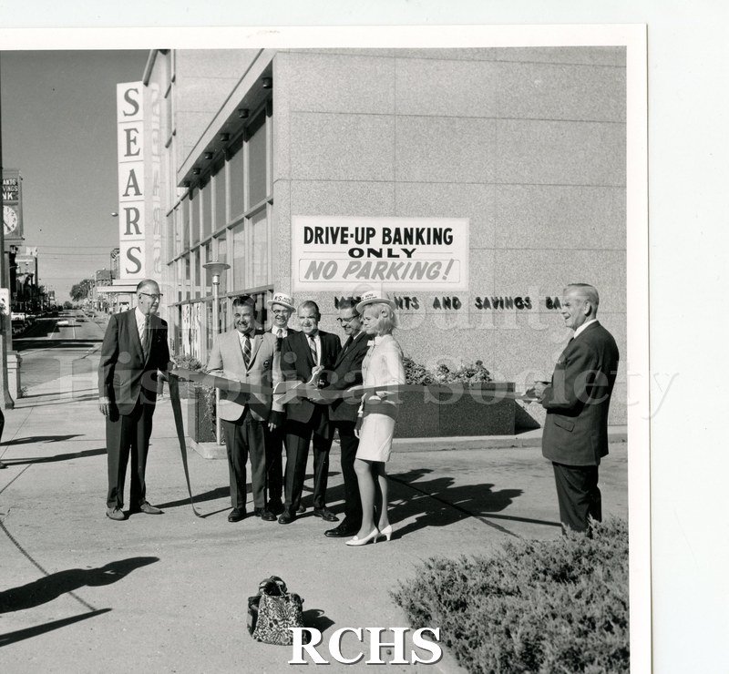 Black and white photograph of the Merchants & Savings Bank ribbon cutting ceremony. There are seven men and one woman holding a ribbon ready for cutting.