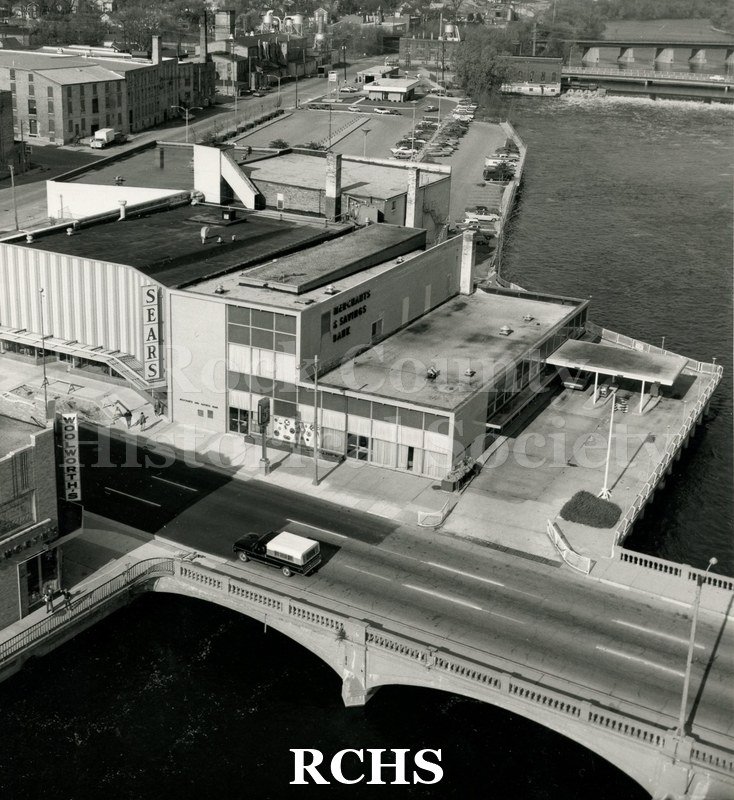 Black and white photograph of the Merchants & Savings Bank from across the river. Shows one vehicle crossing the bridge. There is one car in the drive-thru area.