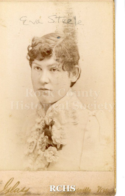 Historic image of Carte-de-visite. Eva Steele. Woman in white dress with lace collar and fischu. Floral arrangement at center, hanging downwards.