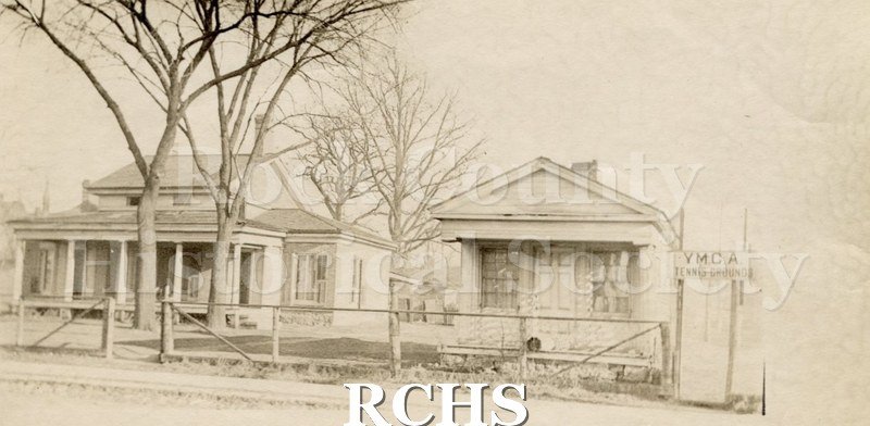 Black and white photograph of YMCA Tennis Grounds, Janesville, WI. Print of a two story house on the left and a smaller building on the right.