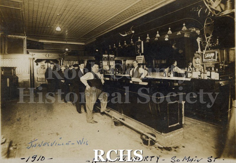 Black and white photograph of Neal Flaherty Bar with a bartender behind the bar and patrons in front. On bottom: "Janesville-Wis 1910 Neal Flaherty-So. Main St.".