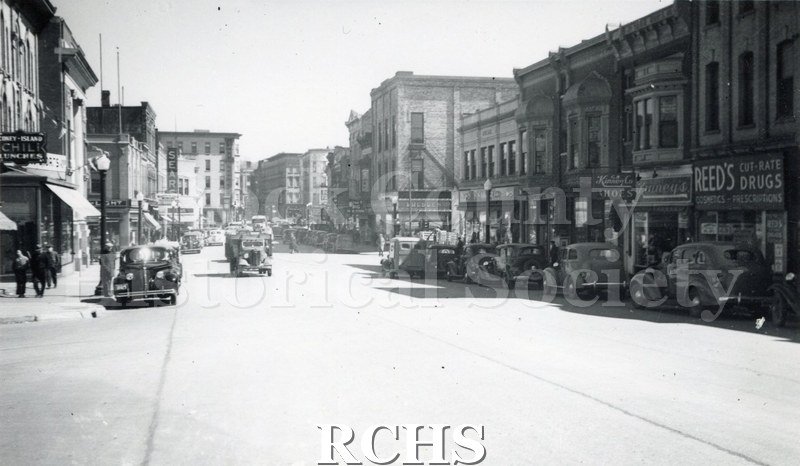 Black and white photograph of Downtown Janesville. On the right is a sign for "Reed's Cut-Rate Drugs" and on the left is the sign for "Sears".