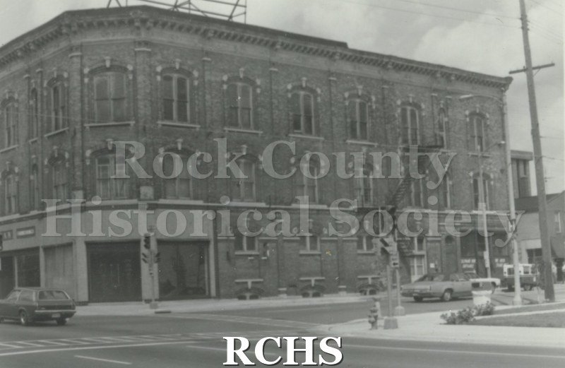 Black and white photograph of the front of an unidentified three story building on a corner of a Janesville street.