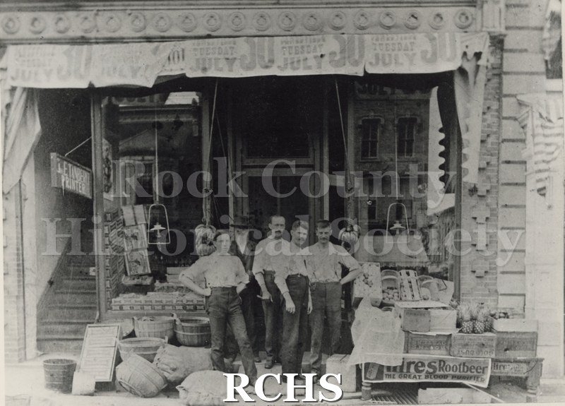 Black and white photograph of an unidentified grocery store on N. Main St., Janesville, WI. There are five unidentified men standing in front of the doorway.