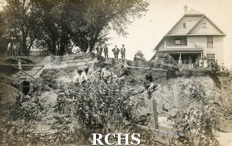 Postcard. Black and white photograph of several men and women surveying a large section of missing road.