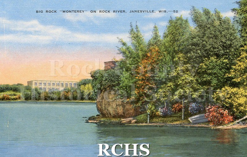 Color postcard. Fall colored trees on shoreline and rock, water in foreground, white building in background.