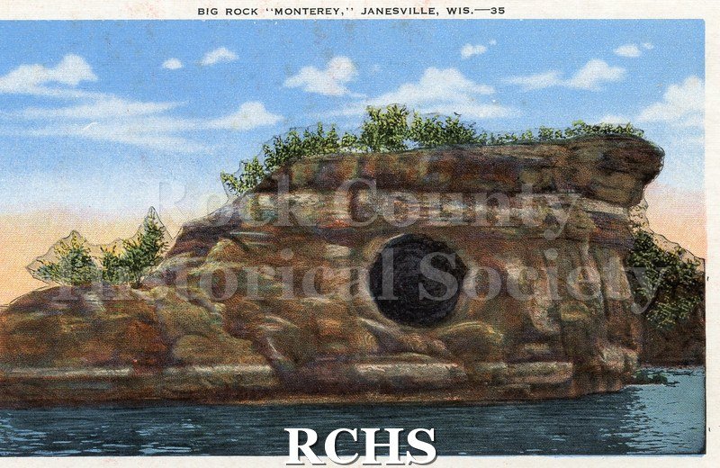 Color postcard. Full color of large rock with round hole in side, trees growing along top, water in front, blue sky with clouds.