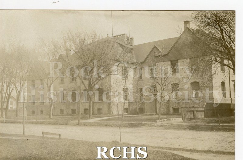 Black and white postcard of building with bare trees, grass and road in foreground. Fence on right with part of house visible. Building is 2 1/2 stories.