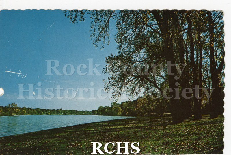 Postcard of Riverside Park and the Rock River. Trees on right and in background. Grass with leaves in foreground. Top and right edges scalloped.