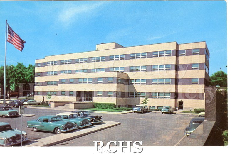 Postcard of Rock County Court House. Flag on and trees on left, cars parked throughout parking lot. Bushes near building. Reverse has no writing, postmark or stamp.