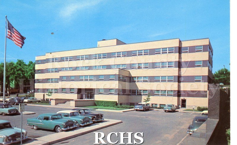 Postcard of Rock County Court House. Flag on and trees on left, cars parked throughout parking lot. Bushes near building. Reverse has no writing, postmark or stamp.