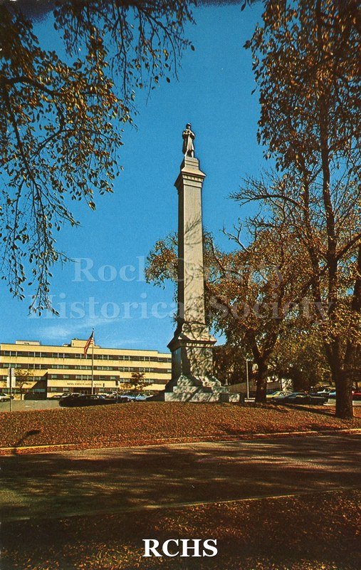 Historic image of Post Card of Court House park. Shows memorial statue with building in background, trees framing edges. All edges scalloped.