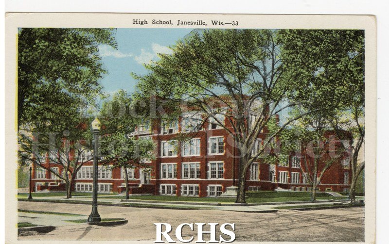 Black and white photograph of Janesville High School. Shows a red brick building with white stone window trim, trees in foreground, corner street, lamp post.