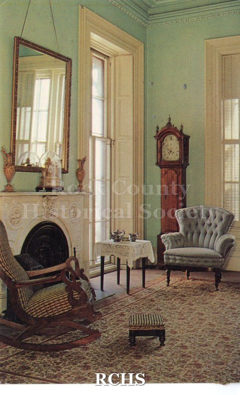 Color postcard of the library of the Lincoln-Tallman House. Blue green painted walls, white modling. One window shutters folded back and othe with louvers open.