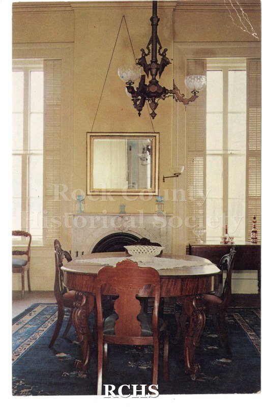 Color postcard of the dining room of the Lincoln-Tallman House. Yellow painted walls, white modling. Both windows partially open and lovenres closed.