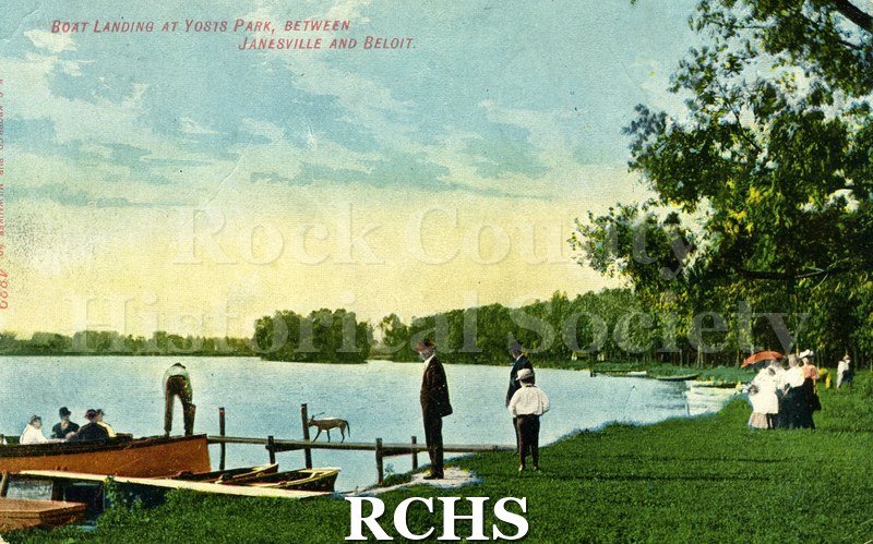 Postcard of boat landing at Yosts park. People in boat on left, car walking across dock, 2 men and a boy standing in center, women with a red parasol on right.