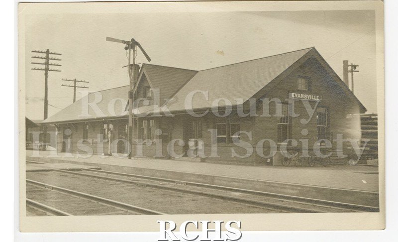 Postcard. Evansville train depot with tracks in front. Sign on end of depot "Evansville". Telephone poles and smokestack in background.