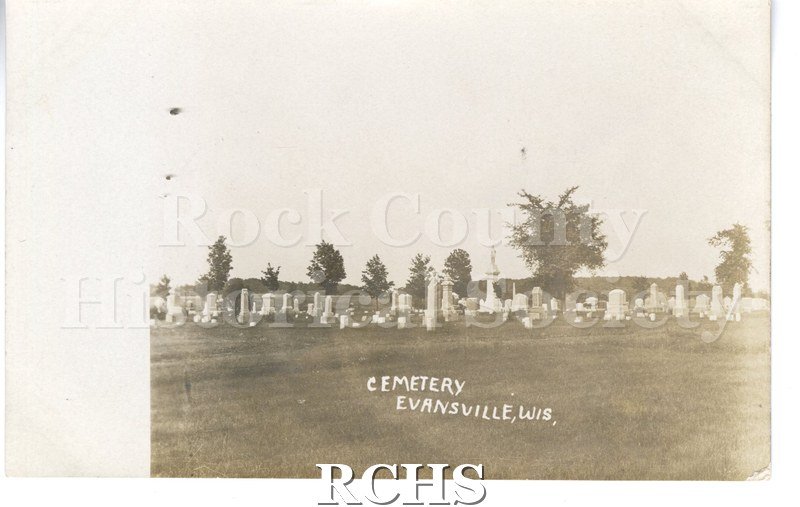 Postcard. Evansville Cemetery. White grave stones with trees scattered throughout. Lawn in foreground. Printed on front in white "Cemetery/Evansville, Wis.