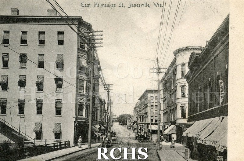 Postcard with black and white picture of East Milwaukee St., Janesville, Wis. Shows horse and wagon on street and people walking on sidewalks.