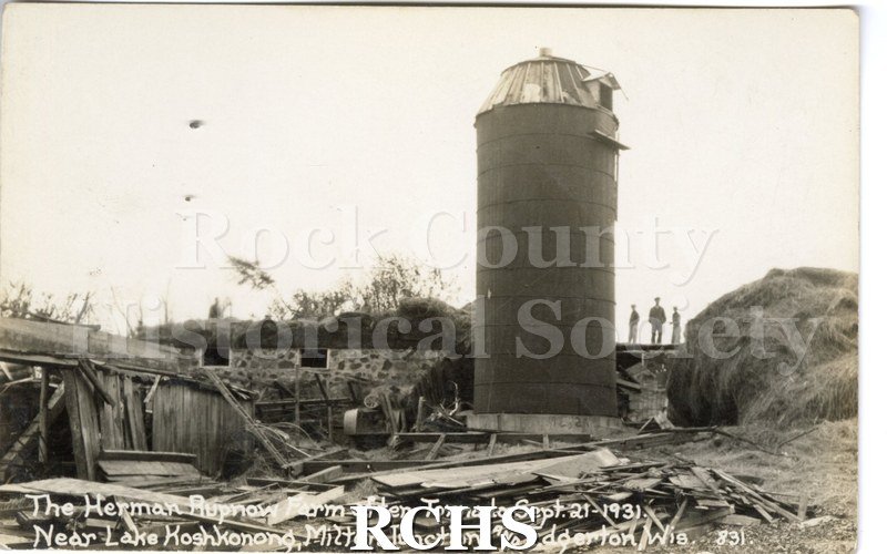 3-1/2" x 5-1/2" glossy picture postcard showing farm after tornado. Silo is visible with three people standing near it. There is a mound of hay on the right side.