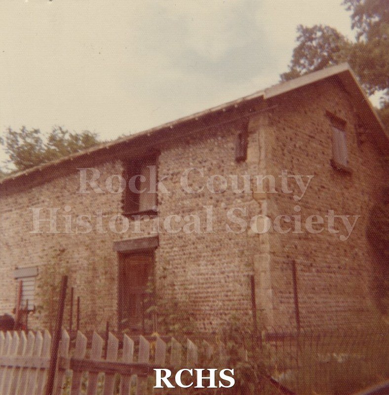 Color photo of the Cobblestone barn behind 910 Broad St., Beloit, WI. Shows a two-story barn with a fence in front of it.