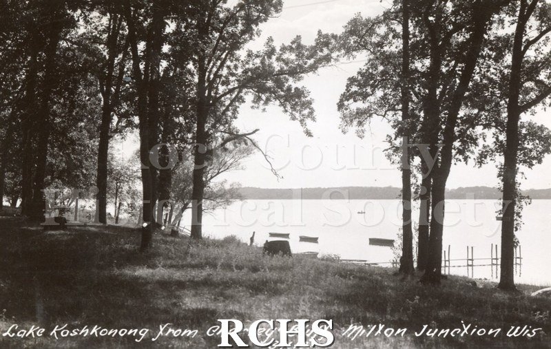 Postcard, photograph of lake scene. "Lake Koshkonong from Charley Bluff. Milton Junction Wis/ 34612". Trees in foreground, small dock visible, boats in water.