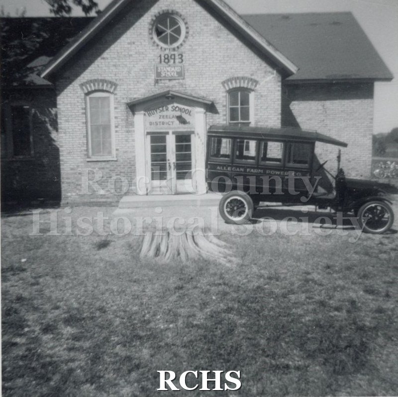 Black and white photograph of the Janesville Carriage Works vehicle at Huyser School. Shows a brick building with a vehicle parked in front of it.
