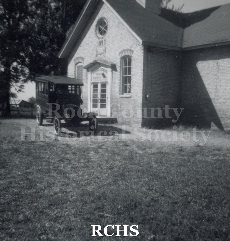 Black and white photograph of the Janesville Carriage Works' vehicle at Huyser School. Shows a brick building with a vehicle parked in front of it.