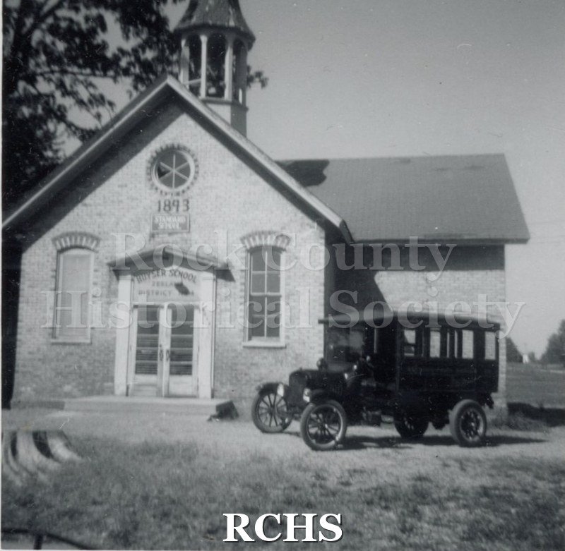 Black and white photograph of the Janesville Carriage Works vehicle at Huyser School. Shows a brick building with a vehicle parked in front of it.
