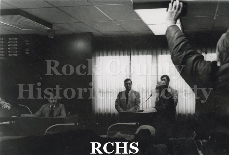 Historic image of Marvin Roth, Tim Cullen, and Gordon Hill are standing at a podium. There are several other people sitting at desks in City Hall.