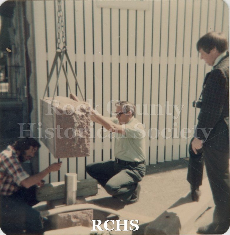 Black and white photograph of two men putting in place a marker using a hoist. Another man is watching on the right.