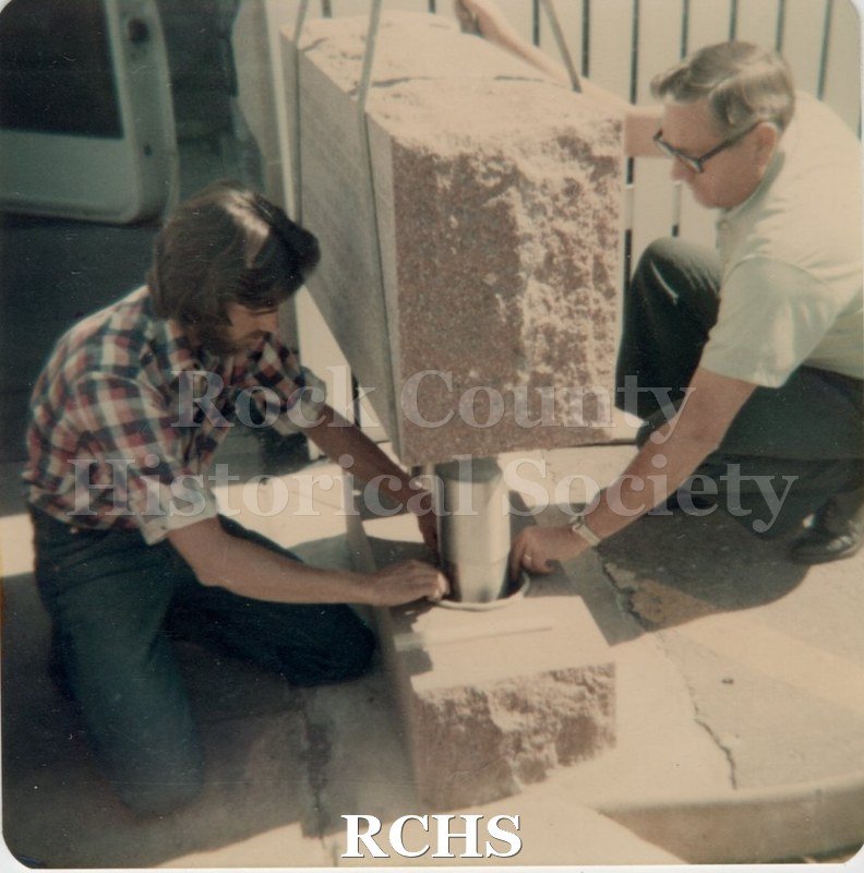 Black and white photograph of two men lowering the capsule into the container. The monument is on a hoist above it.